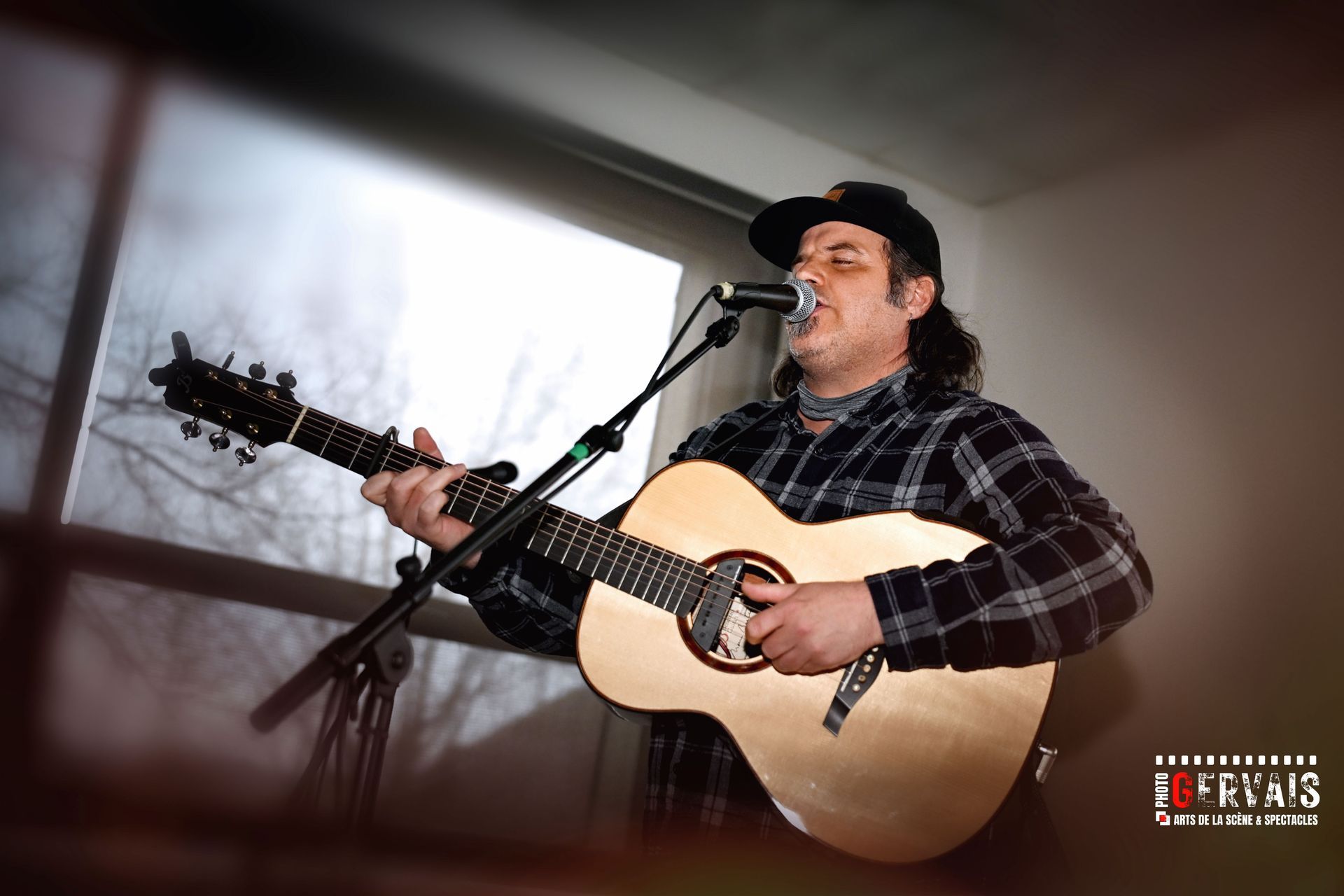 A man wearing a baseball cap wearing a checkered shirt playing a guitar and singing