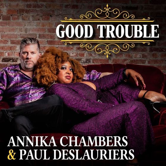 Paul Deslauriers and Annika Chambers sitting on a couch in front of a brick wall on their album cover Good Trouble
