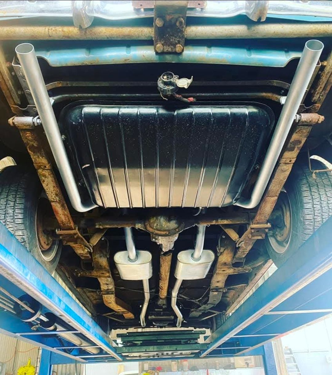Undercarriage of a classic blue car, showing fuel tank, exhaust pipes, and suspension, mounted on a lift.