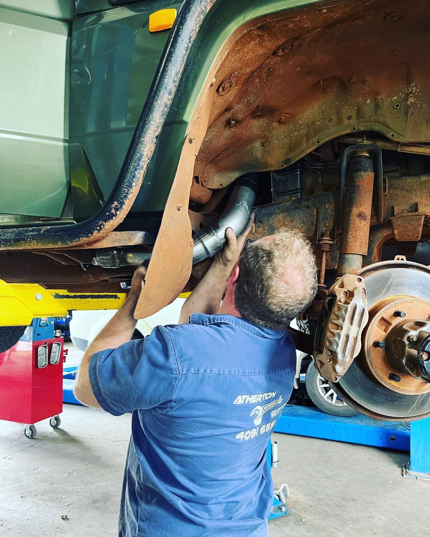 Mechanic Installing Exhaust Pipe on a Lifted Green Vehicle in a Repair Shop — Atherton Mechanical Services in Atherton, QLD