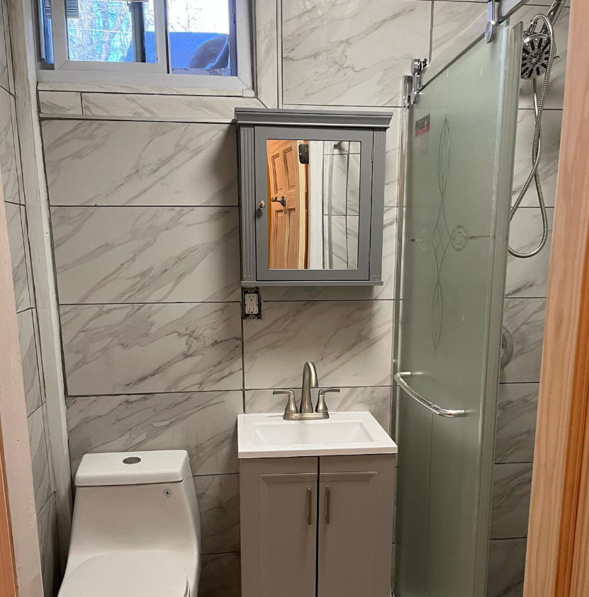 A small bathroom with white marble-patterned tiles, a white toilet, a gray vanity cabinet, and a glass shower stall.