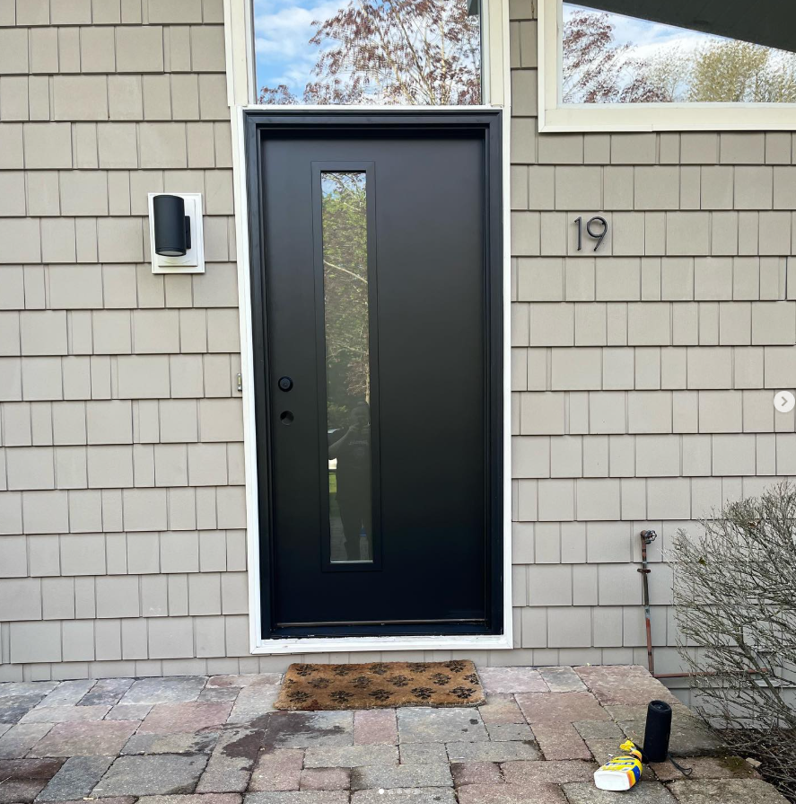 A modern black front door with a vertical glass panel on light-colored shingled siding, with the house number 19 displayed.