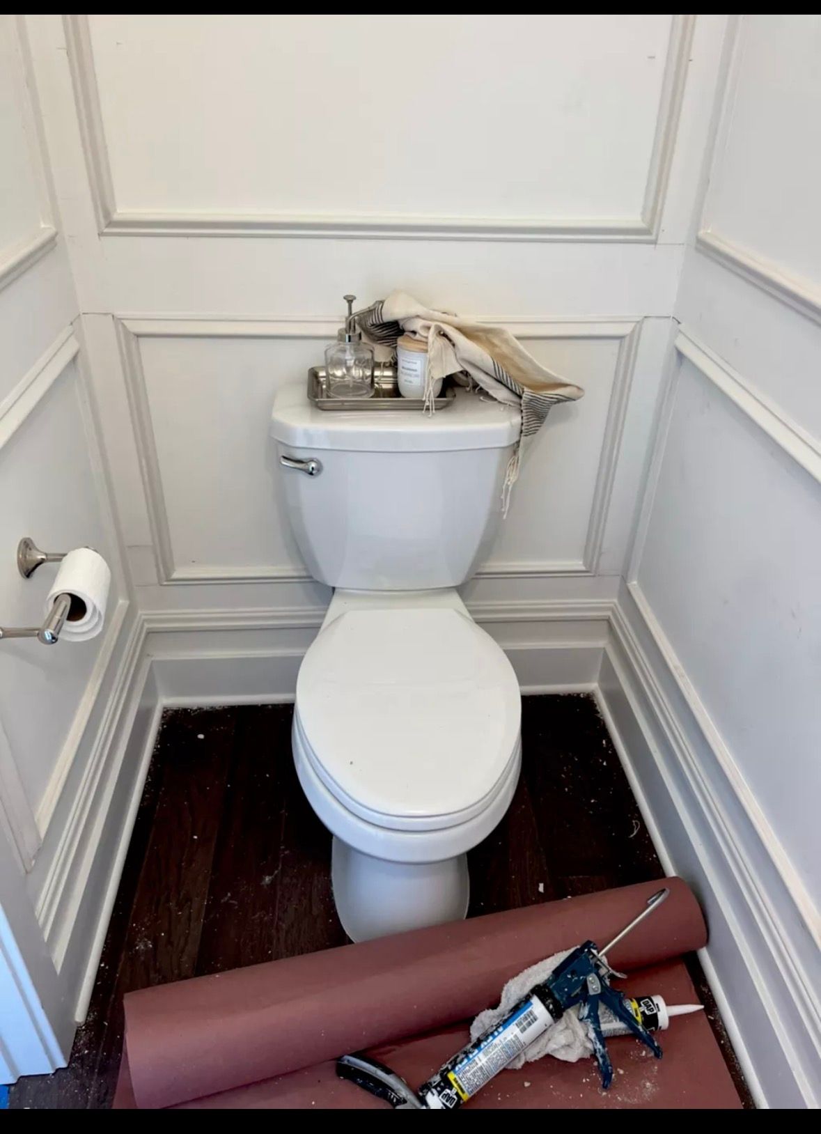 A white toilet stands against a paneled wall in a bathroom, with a caulk gun resting on protective floor paper.