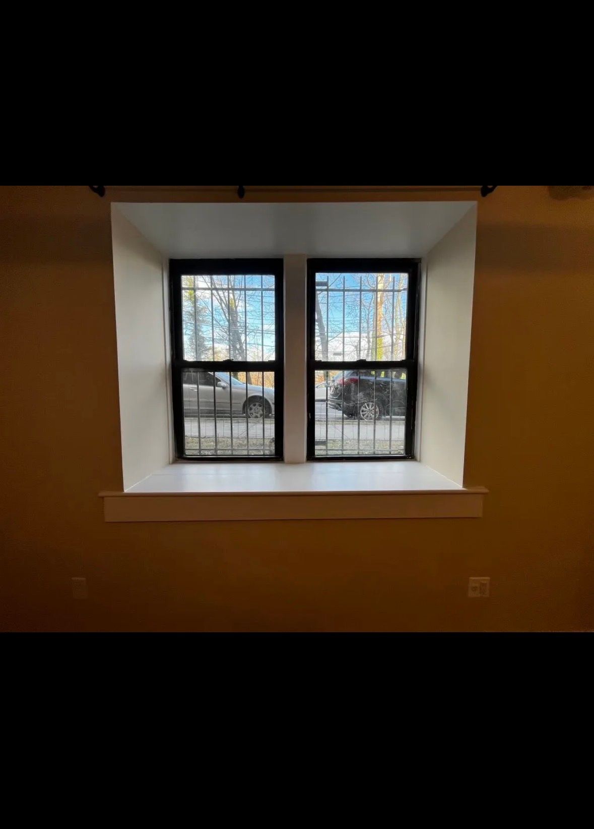 A square window with black frames and security bars looks out onto a street with cars, set in a beige wall with a deep sill.