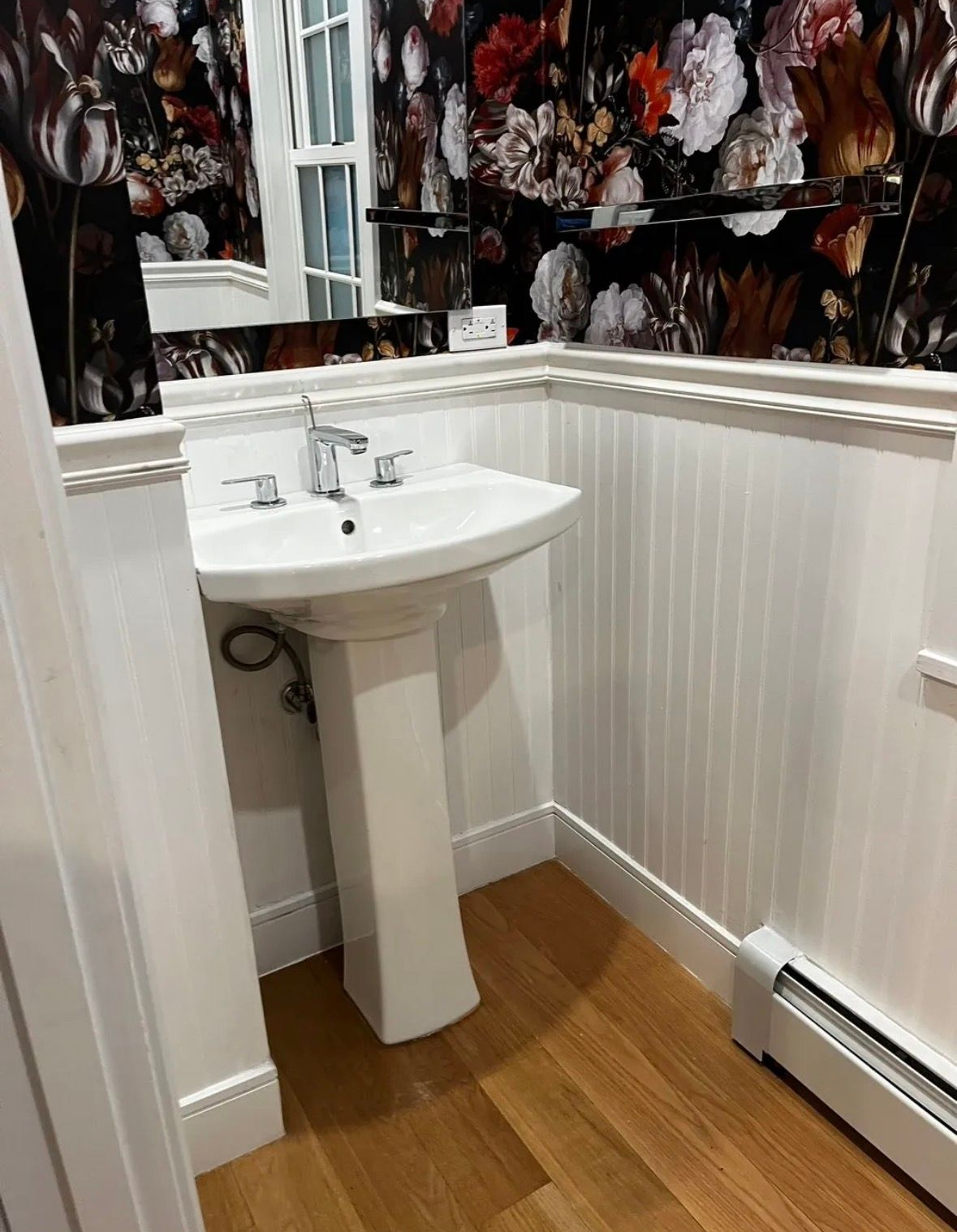 Pedestal sink with chrome faucet against white beadboard paneling and floral wallpaper in a small bathroom.
