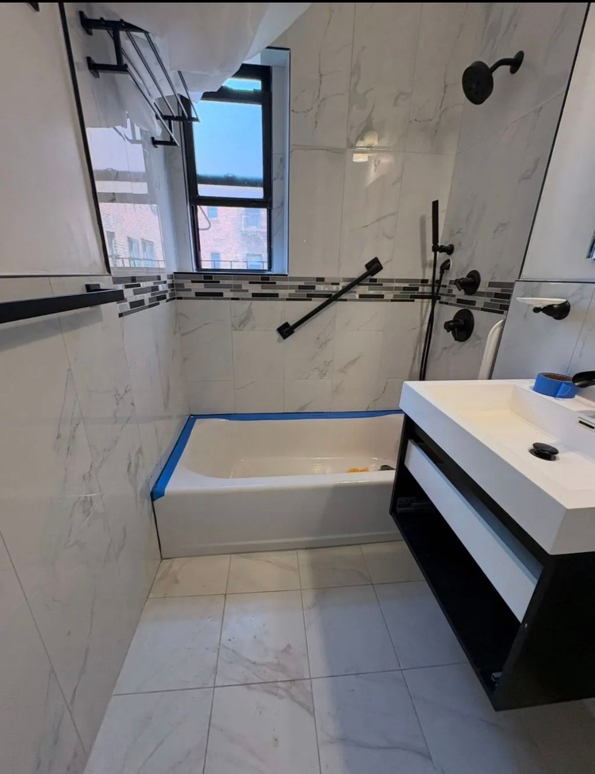 A renovated bathroom featuring white marble-patterned tile, a black vanity, a bathtub with blue painter's tape