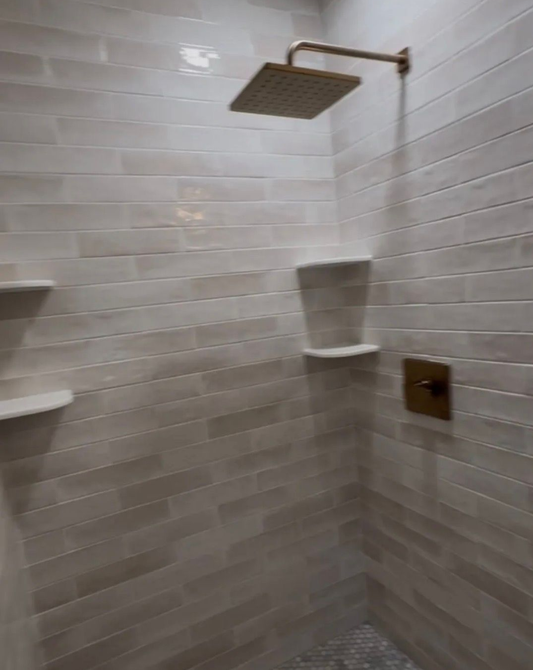 A shower stall featuring light-toned brick-style tiles, a square brass rainfall showerhead, and two built-in corner shelves.