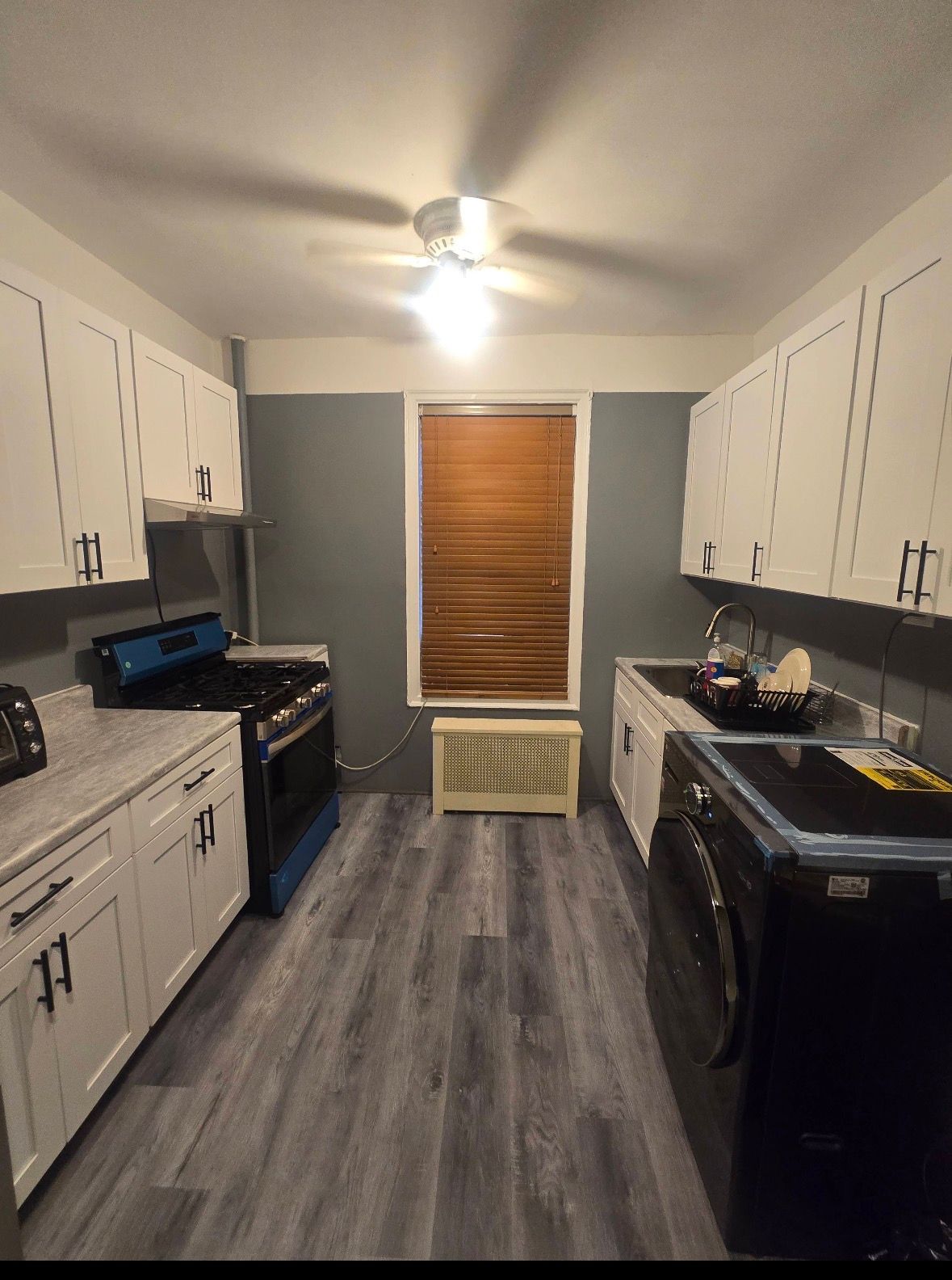 A galley kitchen with white cabinets, gray walls, light-wood flooring, a stove on the left, and a washer on the right.