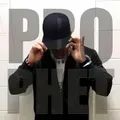 A person wearing a black baseball cap stands in front of a white wall with the text