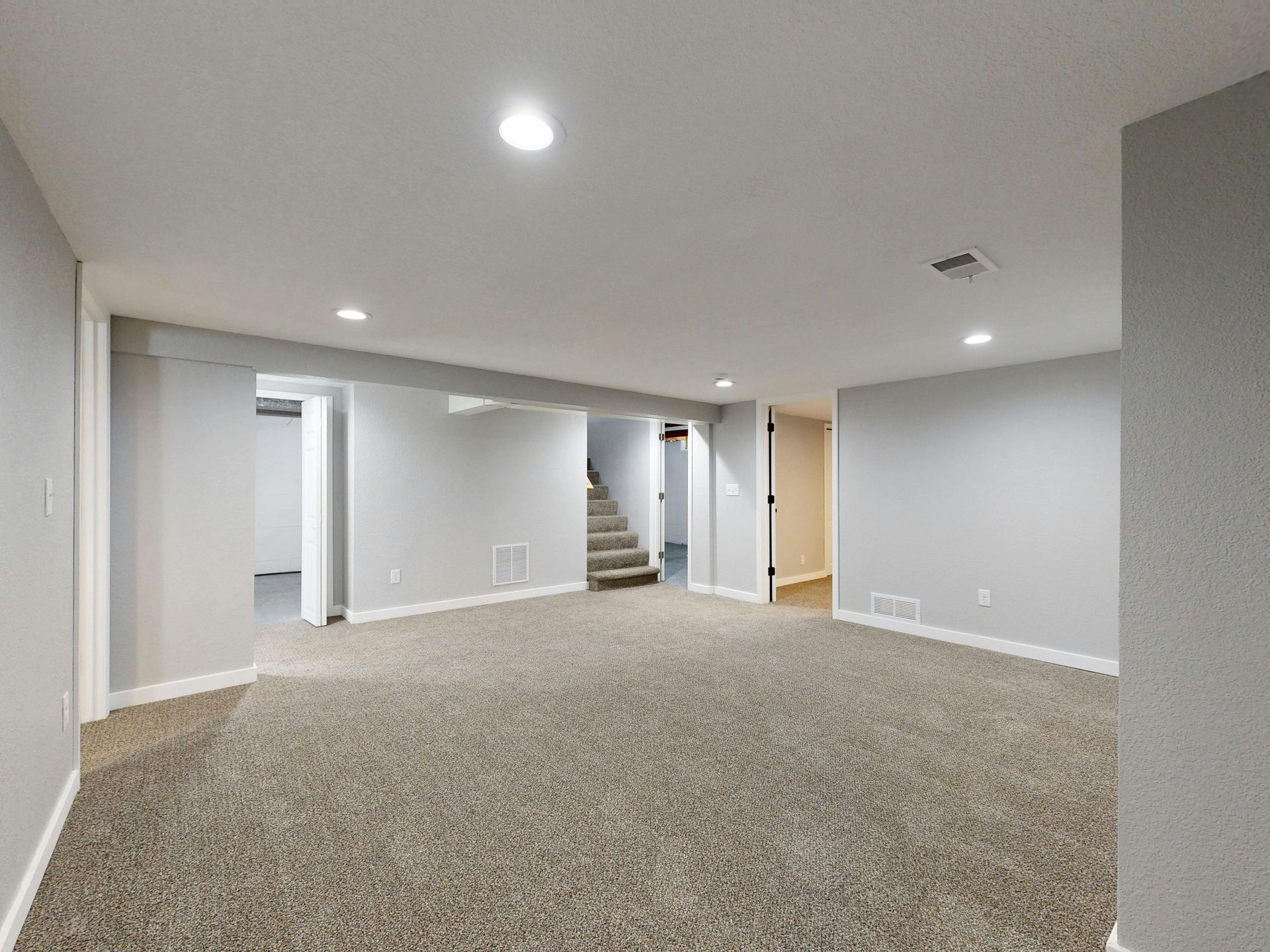 A basement featuring light gray walls, brown carpet, recessed ceiling lights, and stairs leading to the upper level.