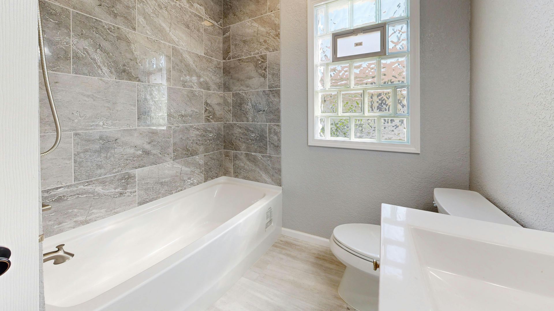 A bathroom with a white bathtub against a grey tiled wall, next to a toilet and a white vanity under a grid window.