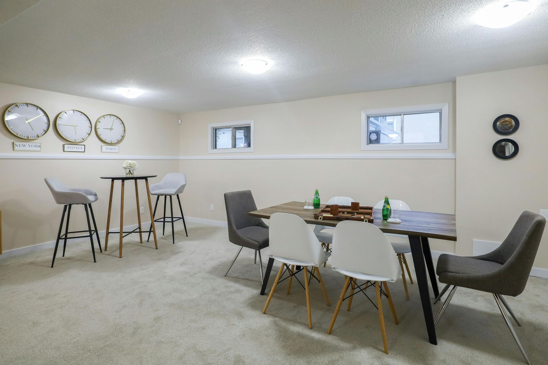 A basement dining area with a wood table, modern chairs, a high-top table, and three decorative clocks on the wall.
