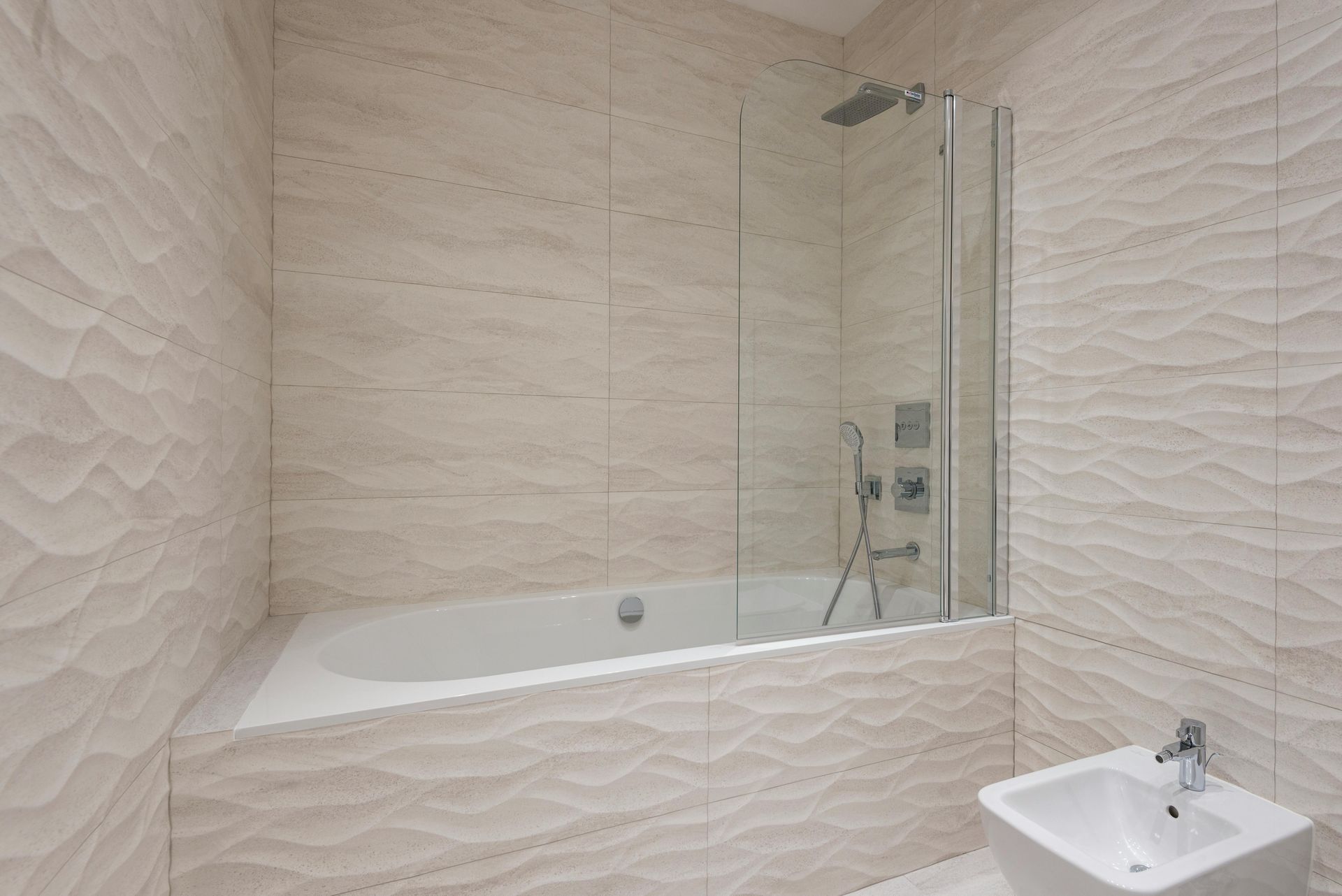 A modern bathroom with textured beige wavy-patterned walls, a white bathtub with a glass shower screen, and a bidet.