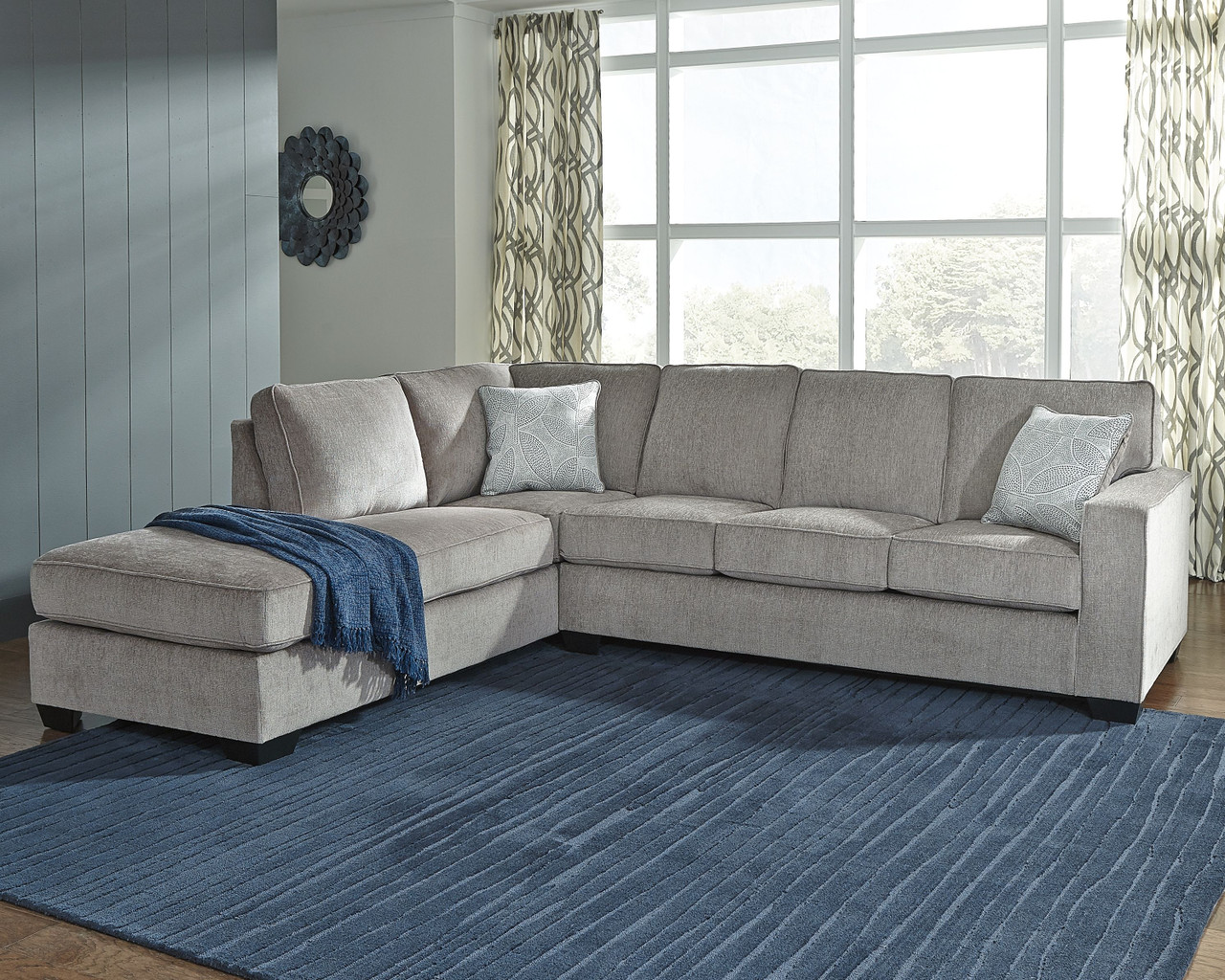 A light gray sectional sofa on a blue rug, with blue and silver throw pillows.