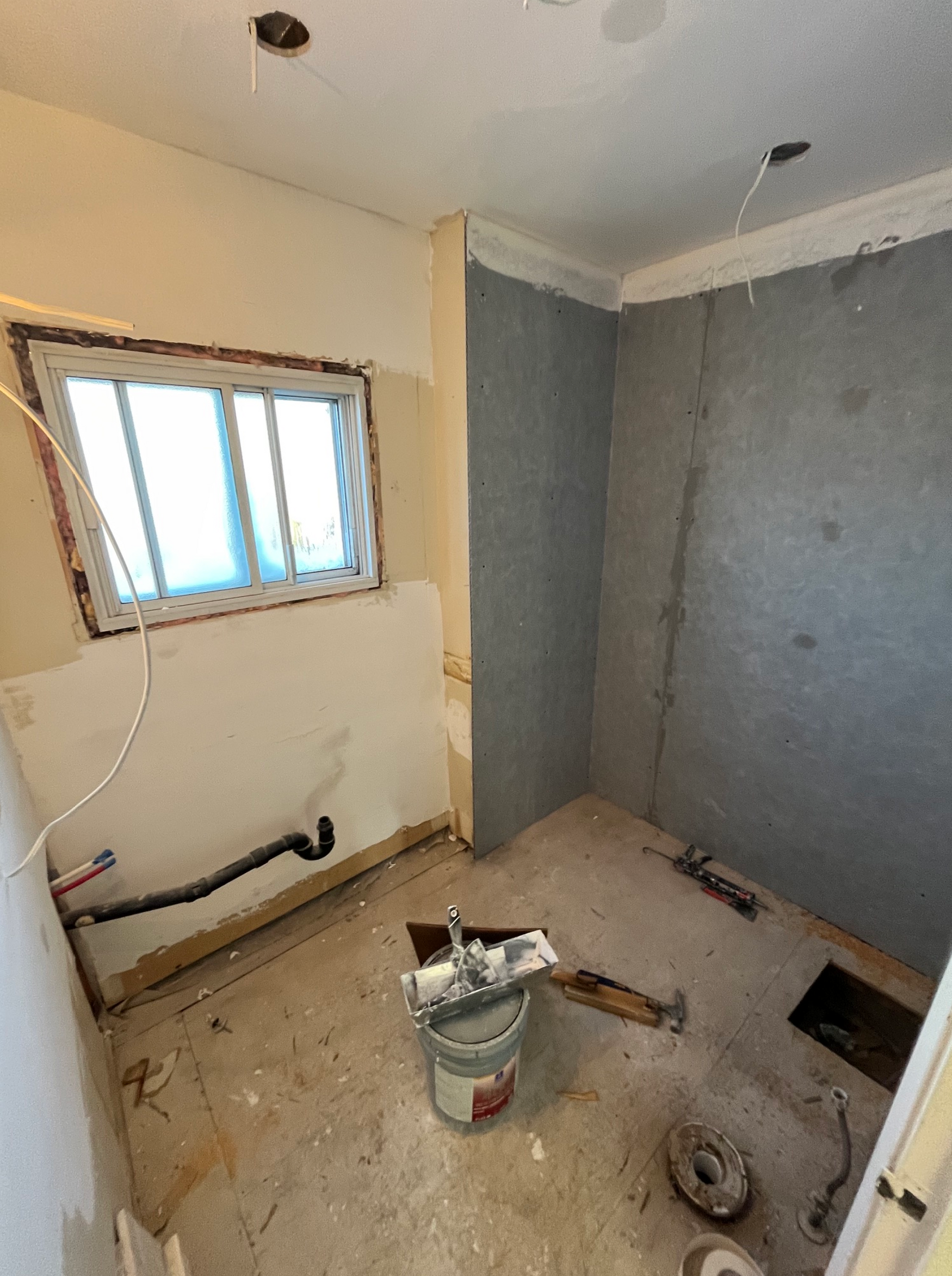 Petite salle de bain inachevée avec des murs gris, une fenêtre, des tuyaux apparents et un seau d'outils posé au sol.