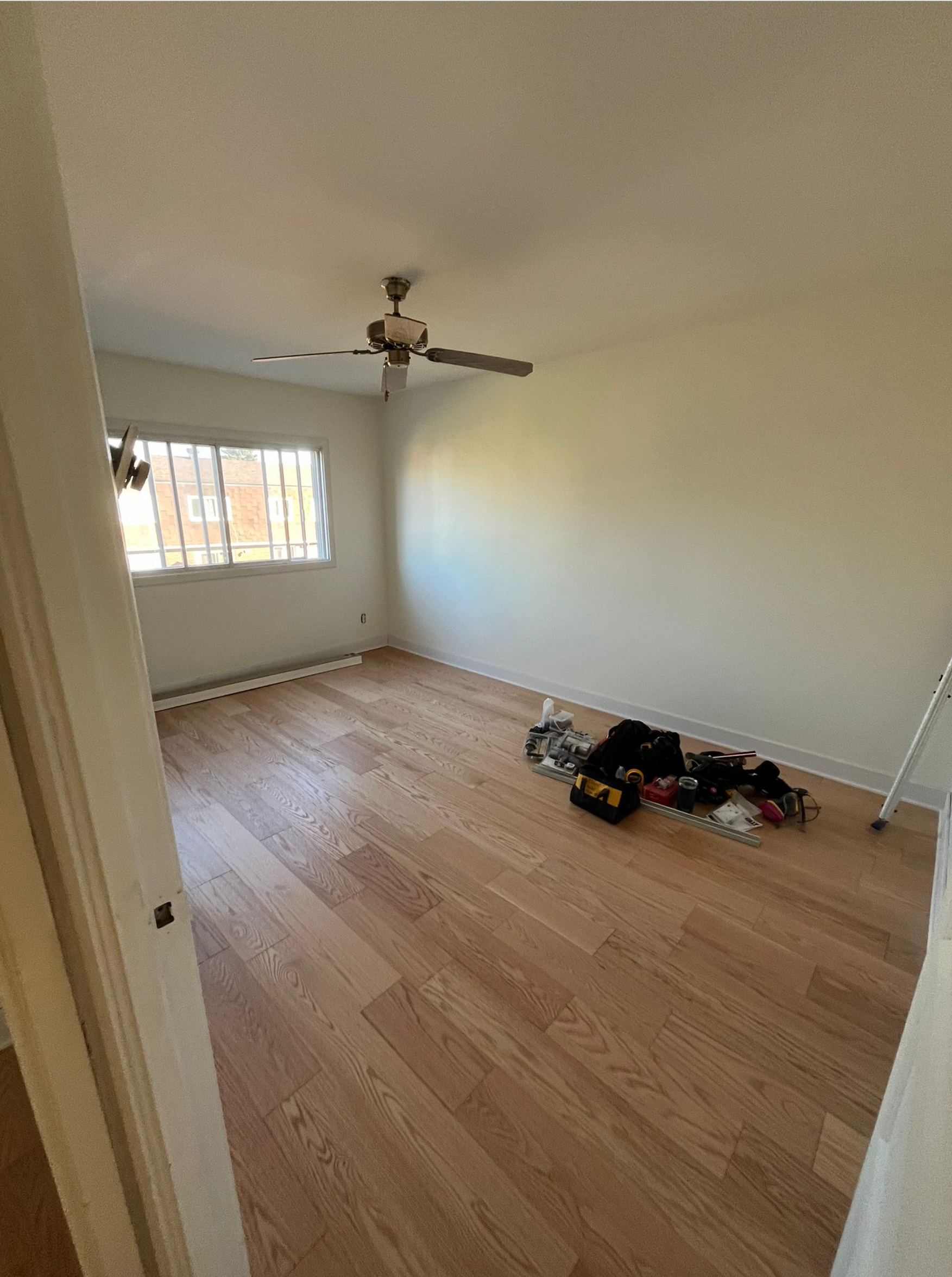 Pièce beige vide avec parquet, ventilateur de plafond et fenêtre ; vêtements empilés sur le sol.