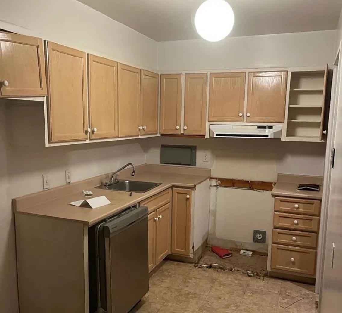 Petite cuisine avec placards en bois clair, plan de travail marron, évier, lave-vaisselle et coin cuisson manquant
