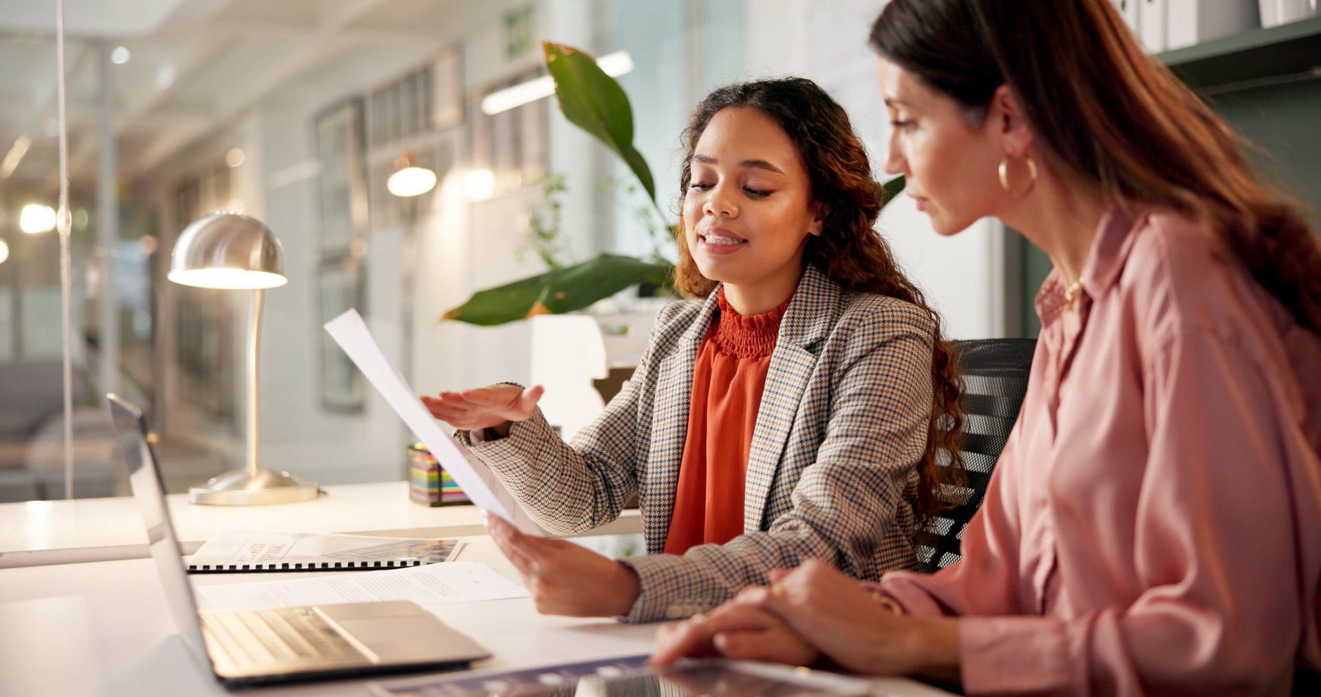 Two people in a modern office review a document together, with one pointing to the page while the other looks on.