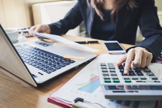 A person in a professional blazer uses a calculator and laptop at a desk with financial charts and a smartphone.