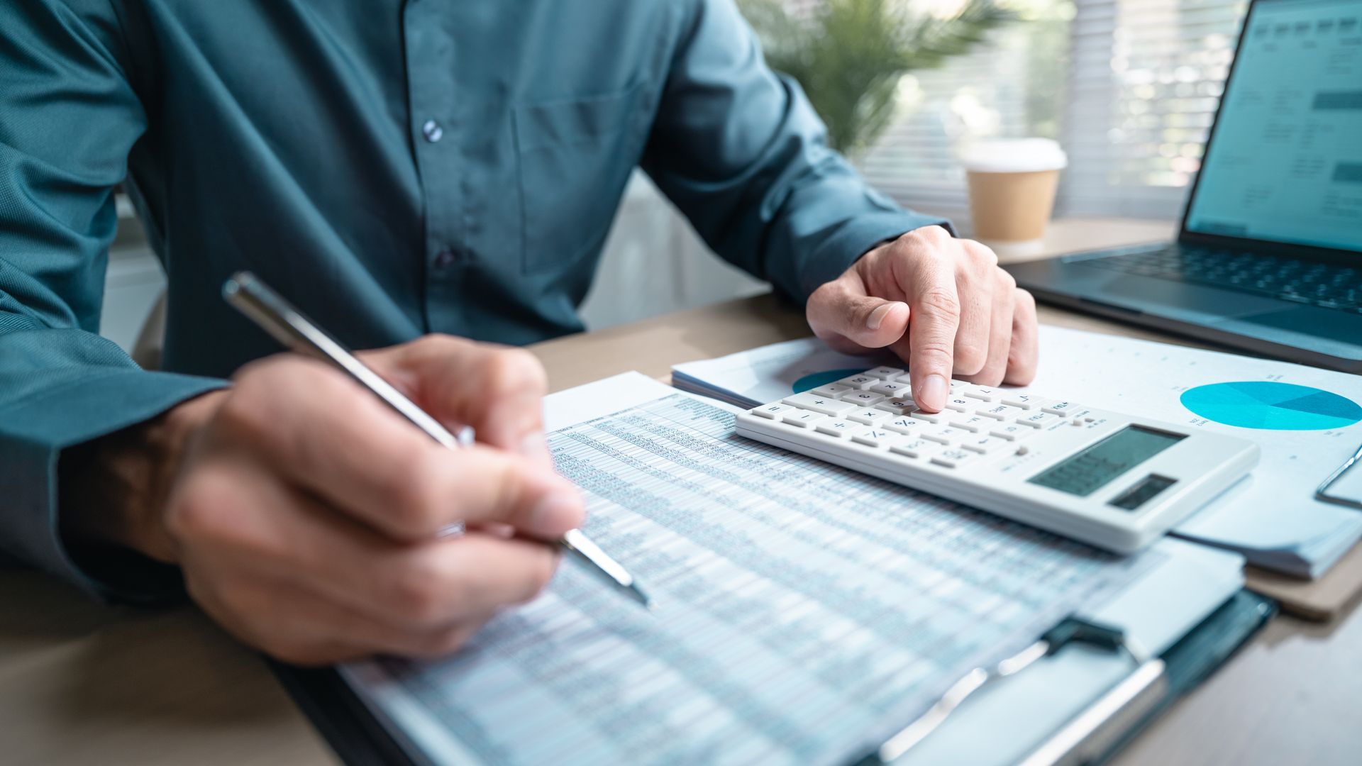 A person in a dark green shirt works at a desk, calculating figures on a paper spreadsheet with a calculator and pen.