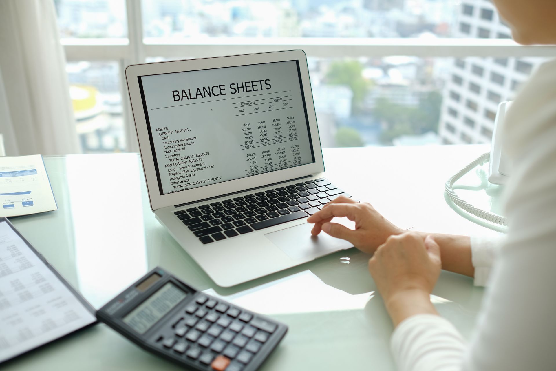 Person working on a laptop displaying a balance sheet at a desk with a calculator.