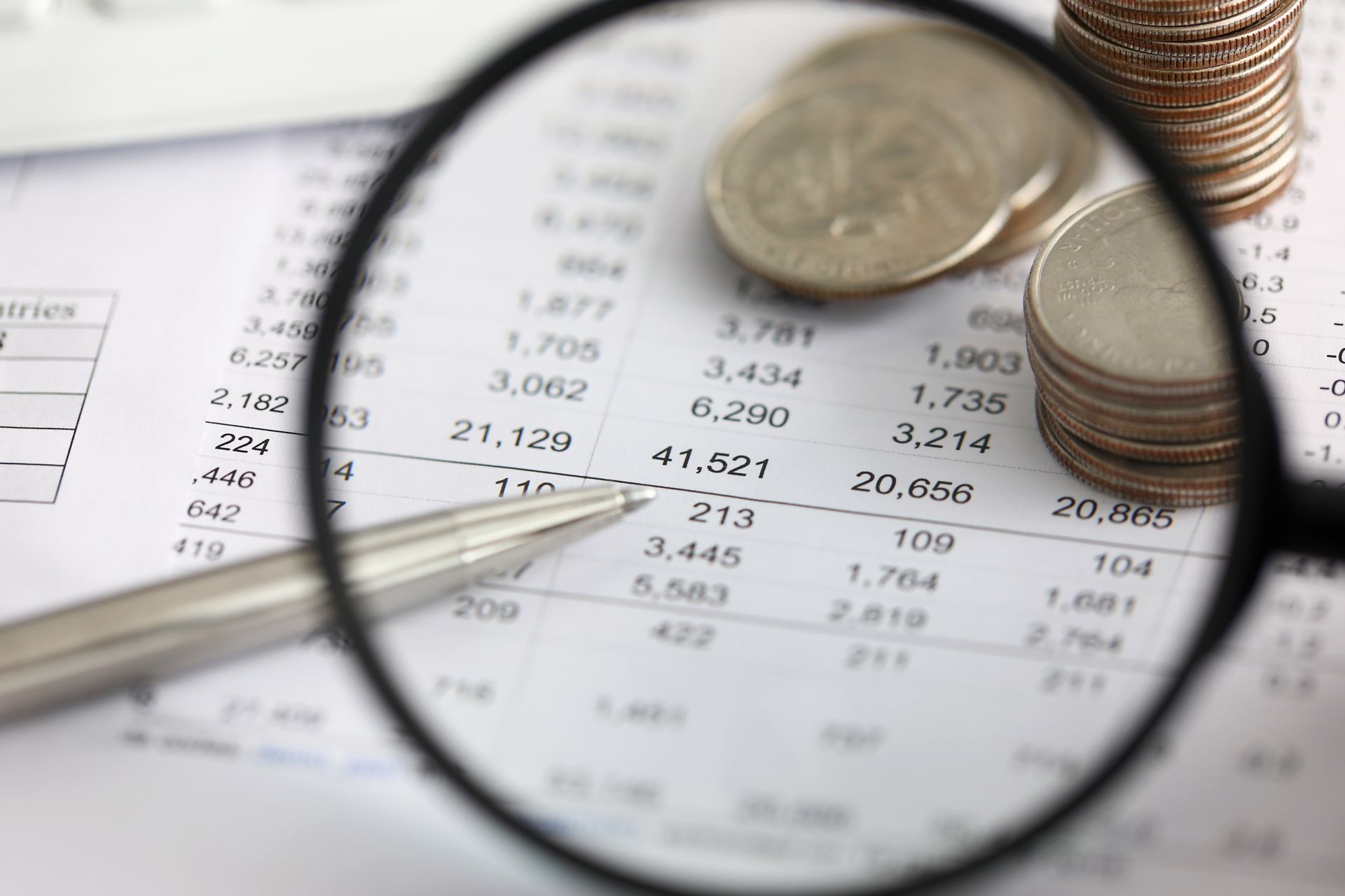 A silver pen and stacks of coins rest on a spreadsheet, viewed through a magnifying glass.