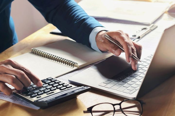 A professional works at a desk, using a calculator and laptop while holding a pen over a notebook.