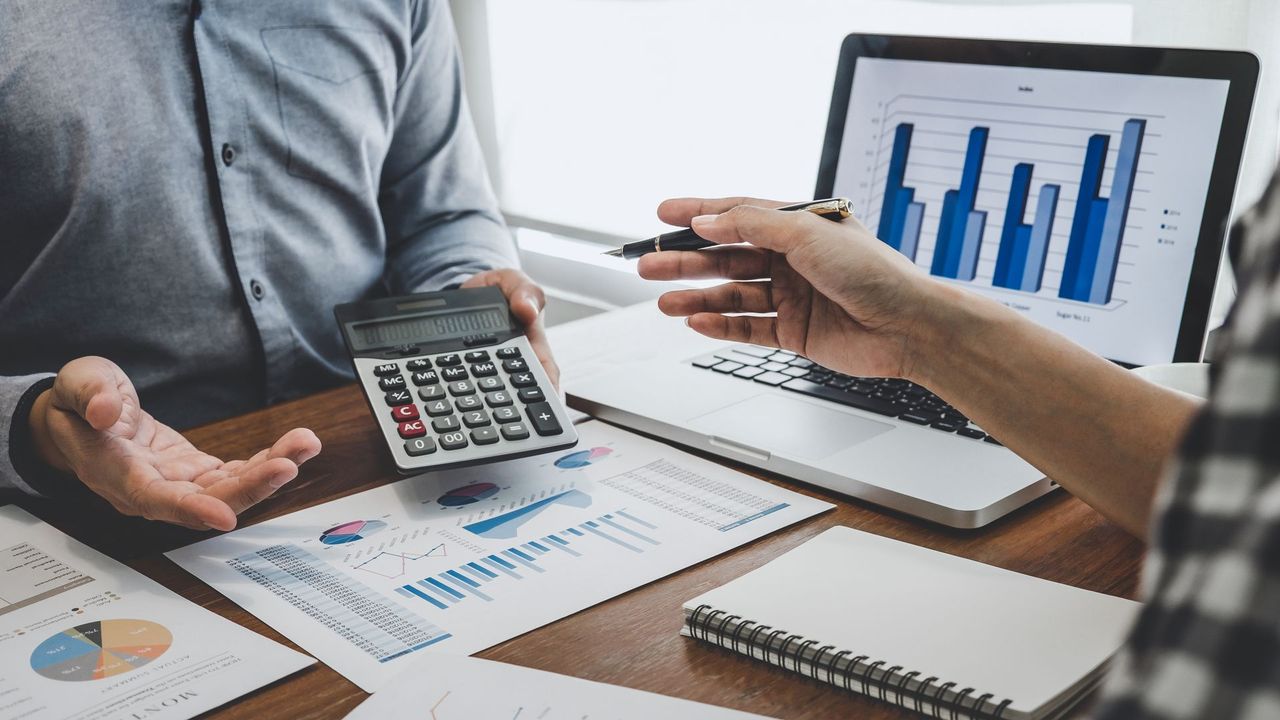 Two people analyze financial charts and graphs on papers and a laptop, using a calculator during a professional meeting.