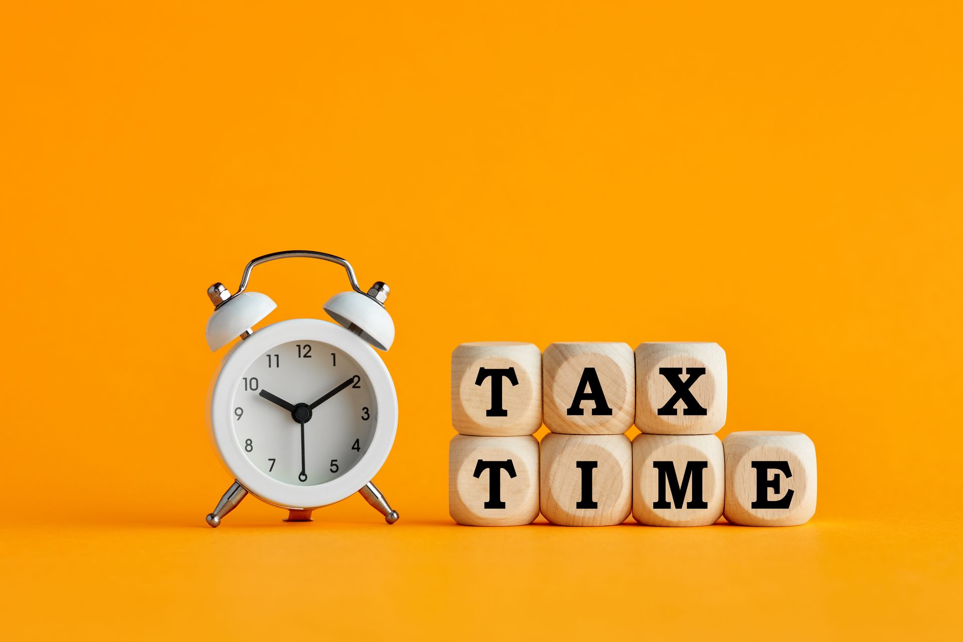 A white alarm clock beside wooden blocks spelling 'TAX TIME' against a bright orange background.
