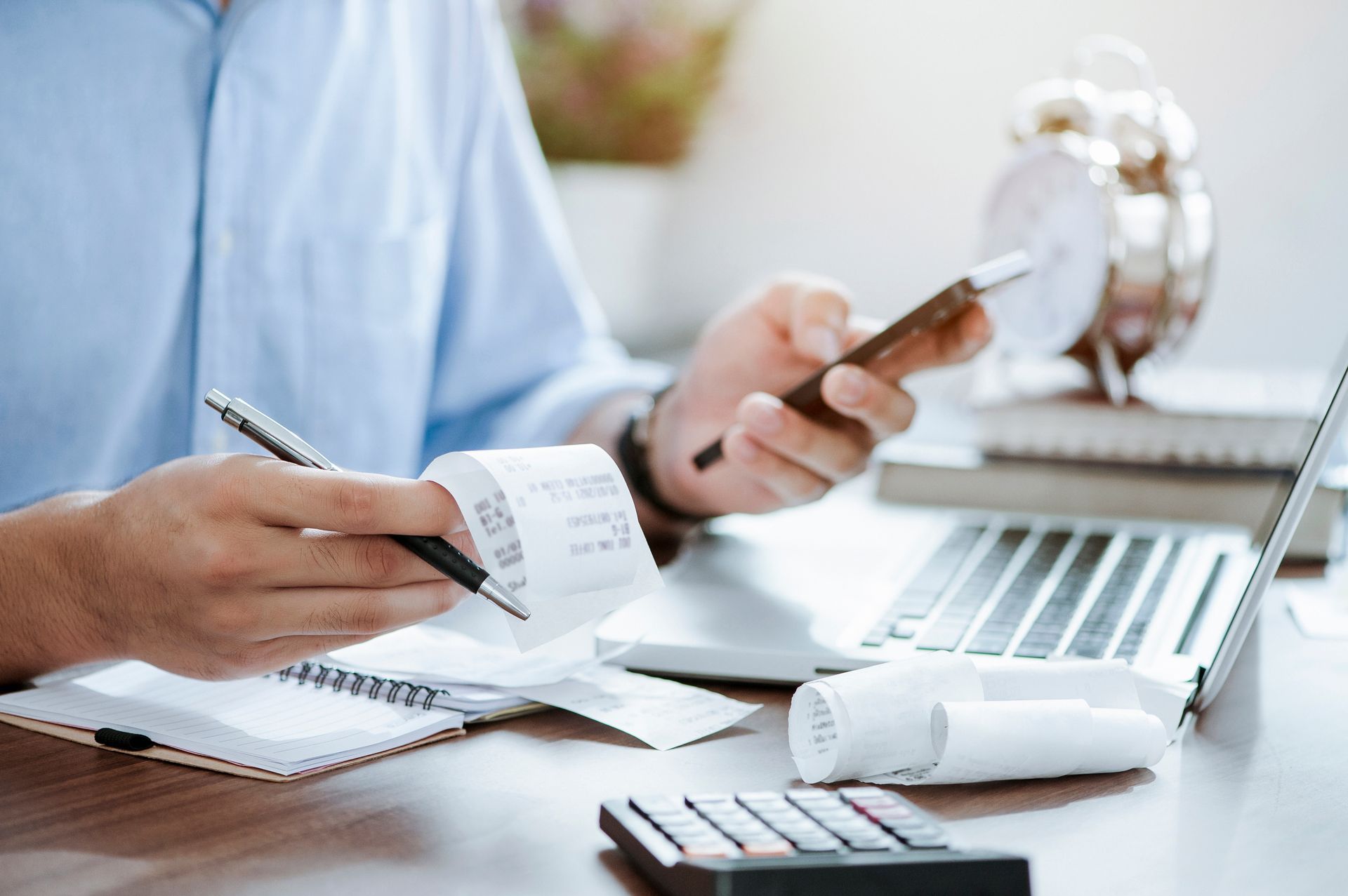 A person in a light blue shirt checking receipts with a pen, a phone, and a laptop on a wooden desk.