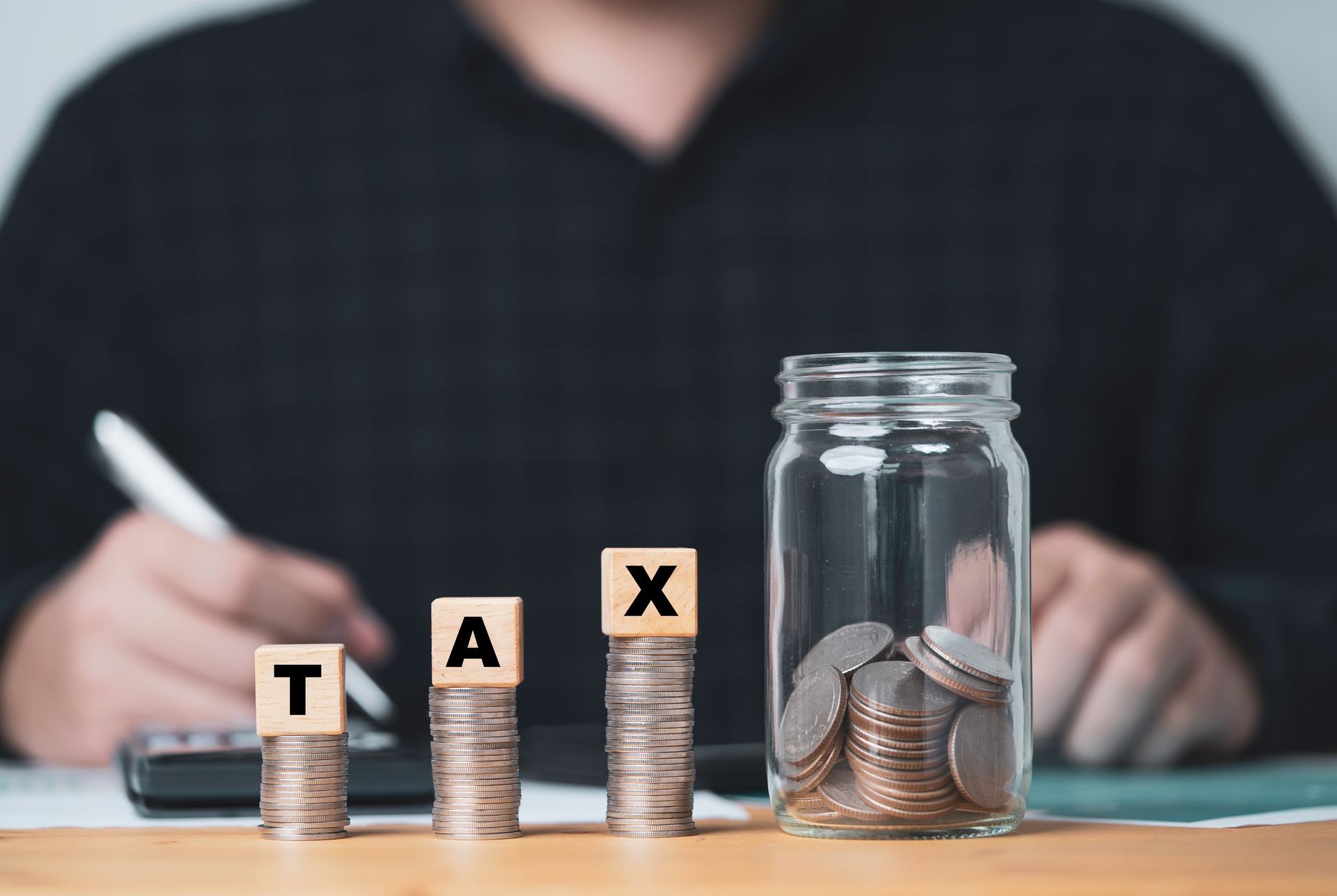 Wooden blocks spelling 'TAX' sit atop three stacks of coins beside a glass jar full of coins, with a person writing nearby.