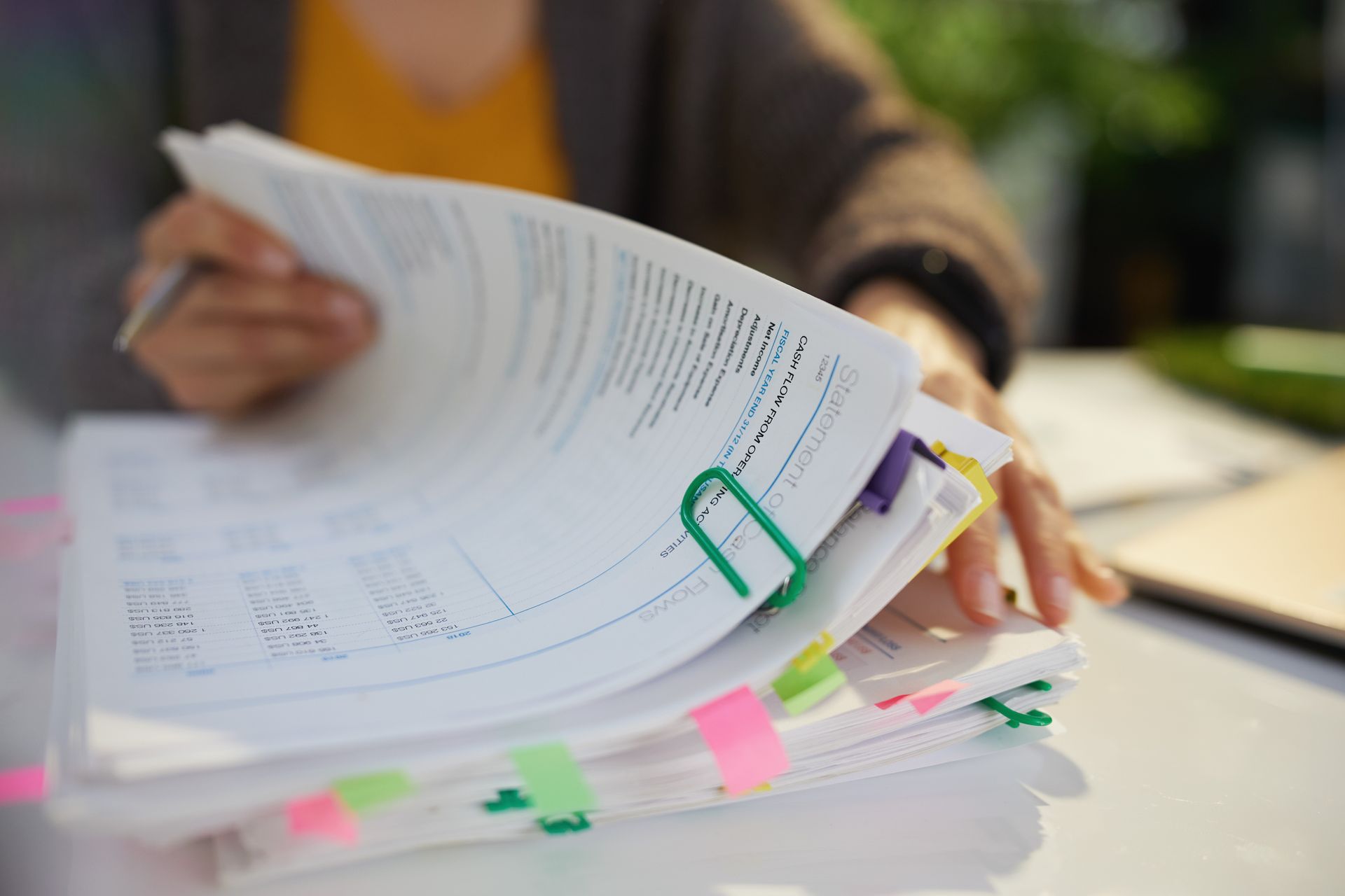 A person's hands turning the pages of a thick document held together by a green clip and marked with colorful tabs. A person's hands turning the pages of a thick document held together by a green clip and marked with colorful tabs.