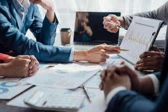 A business team gathers around a table, analyzing charts and data on documents and a laptop during a meeting.