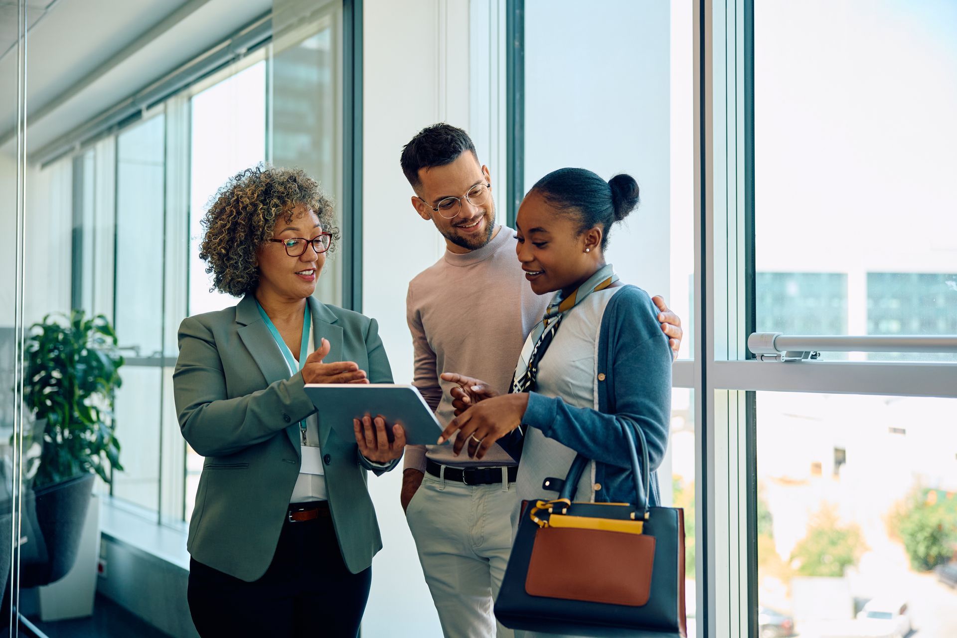 A professional in a blazer shows a tablet to two people in a bright, modern office near a large window.