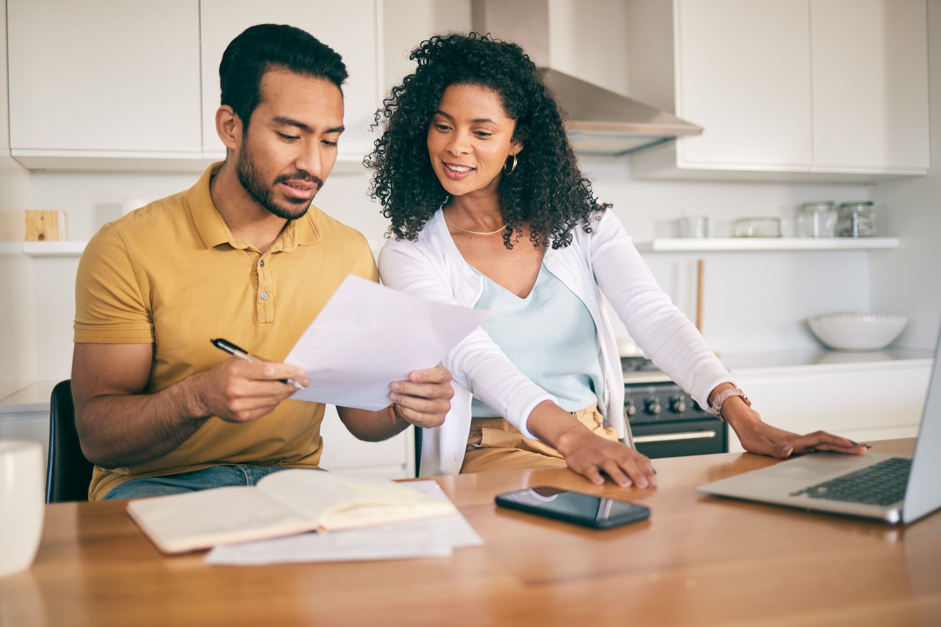 Two people sit at a kitchen table reviewing documents and using a laptop, working together on home finances.