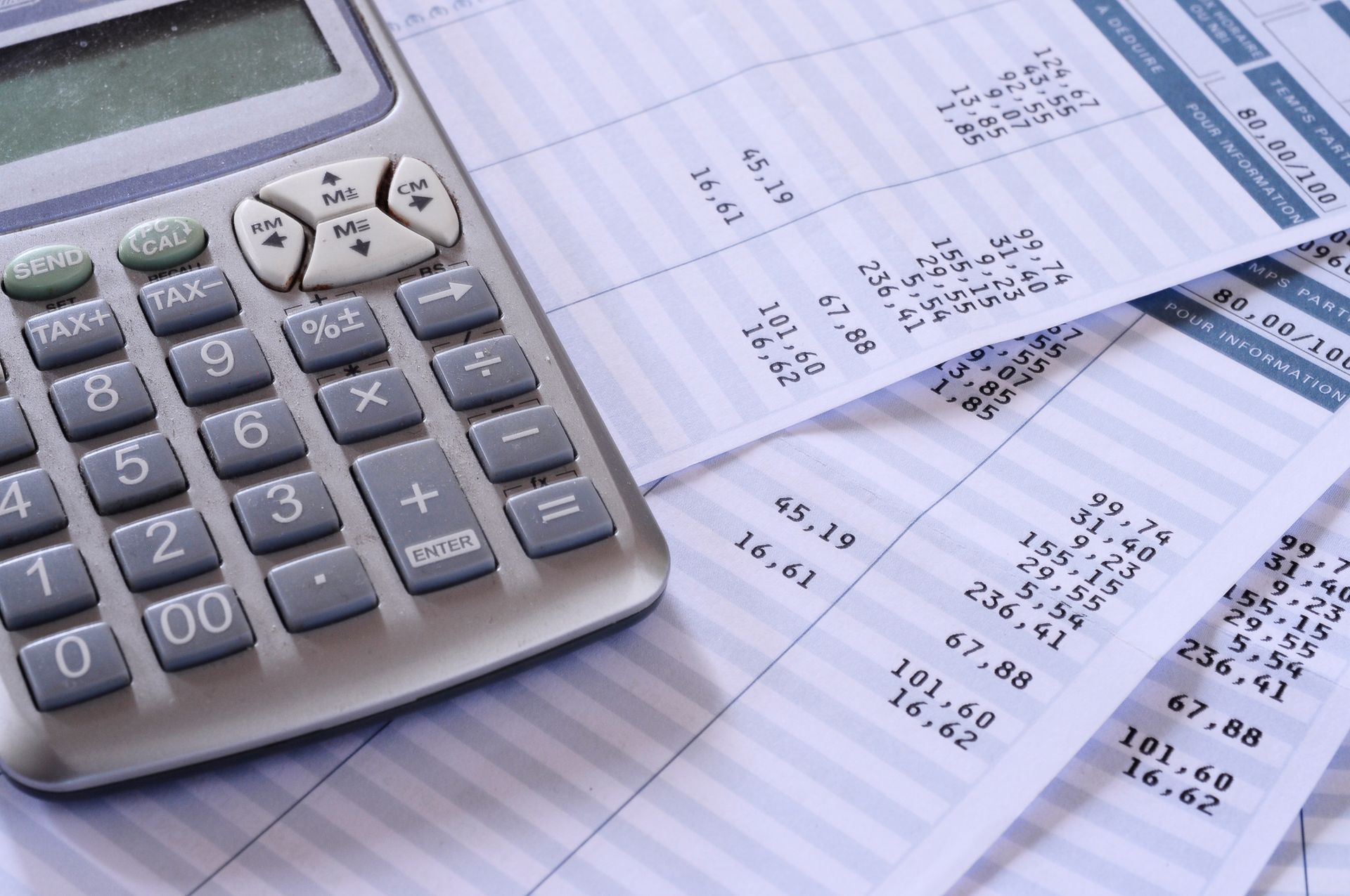 A gray calculator sits on top of several white paper receipts with printed numerical lists and tax calculations.