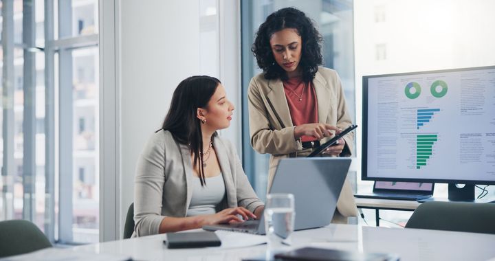 Two people collaborate in a brightly lit office, looking at data displayed on a laptop and a large monitor.