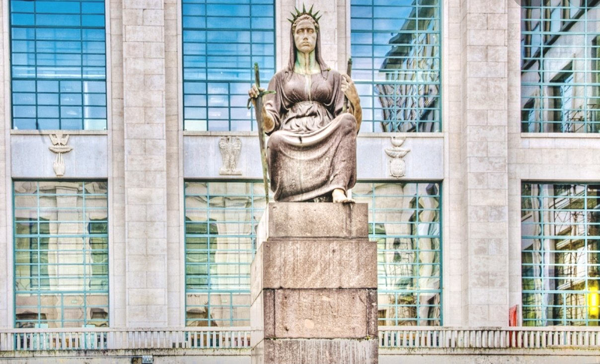 Statua di una donna seduta che tiene in mano un bastone, di fronte a un edificio con grandi finestre.