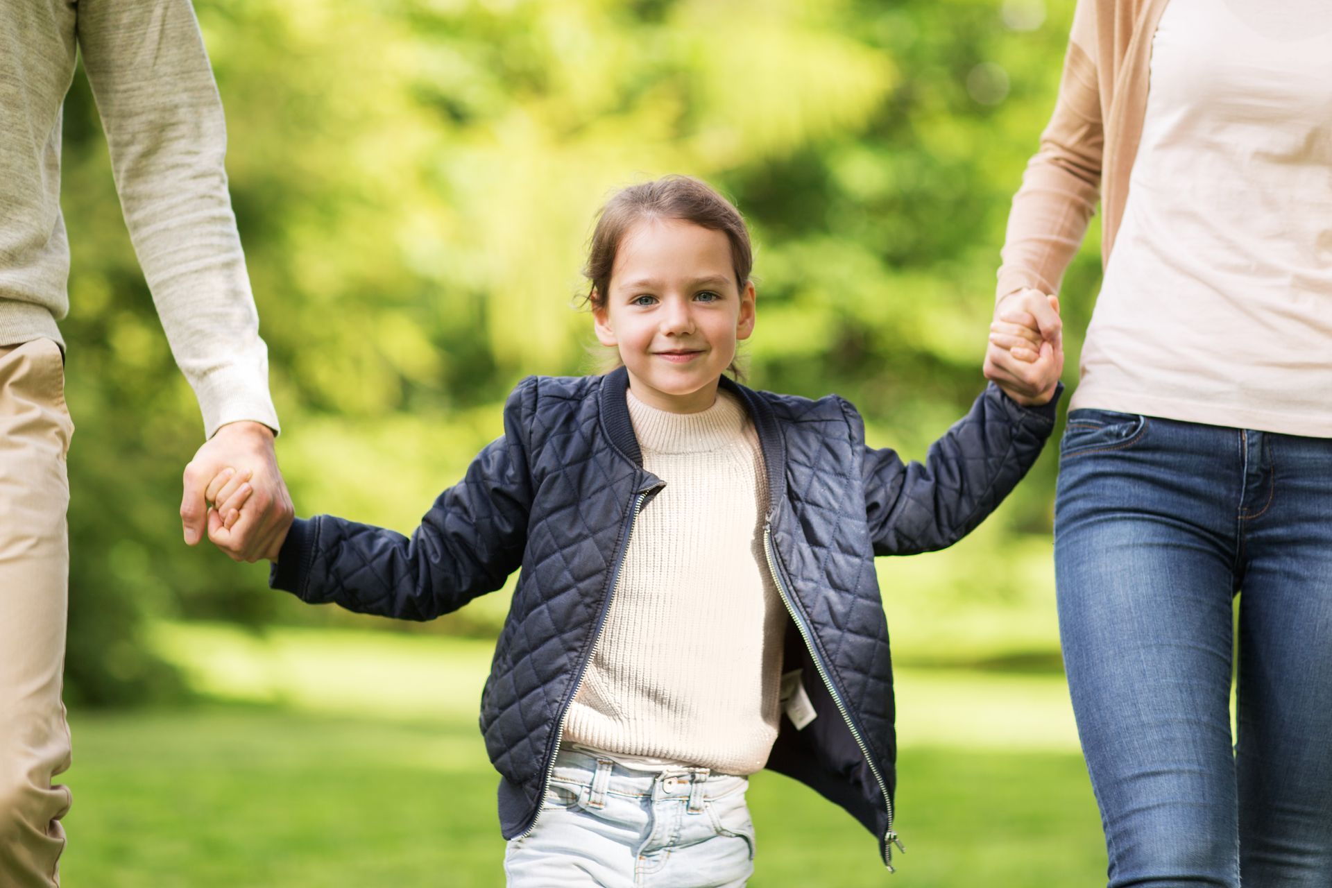 Smiling child holding hands with two adults while walking outdoors in a green park. Smiling child holding hands with two adults while walking outdoors in a green park.