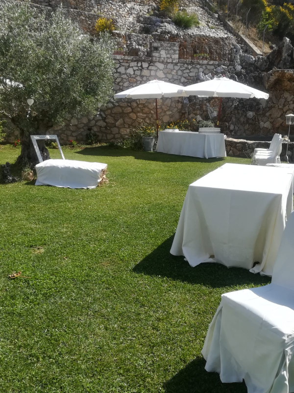 Area esterna con posti a sedere, disposta su un prato, con tavoli e sedie coperti da tovaglie bianche e ombreggiati da ombrelloni, accanto a un muro in pietra.