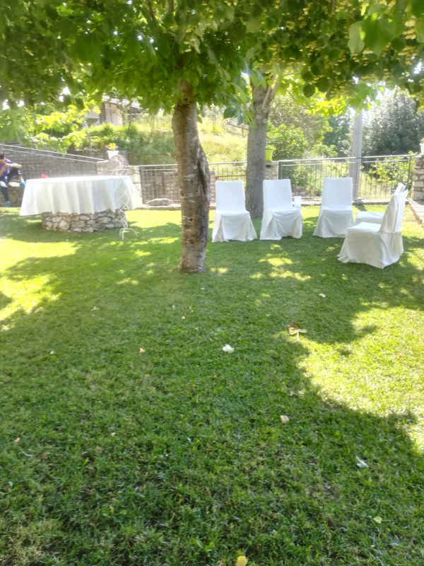Ricevimento in giardino ombreggiato con tavolo rotondo e sedie bianche su un prato erboso sotto gli alberi.