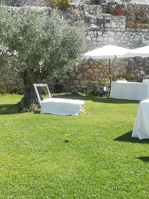 Prato del giardino con sedie e tavoli bianchi sotto ombrelloni accanto a un muro di pietra e un ulivo.
