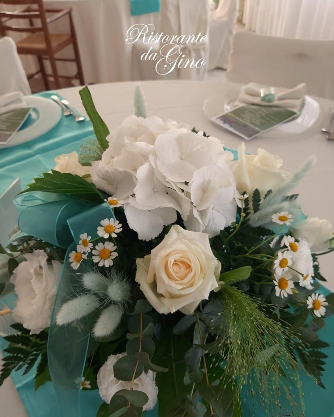 Centrotavola floreale bianco e verde acqua su un tavolo da banchetto apparecchiato, con uno sfondo di ristorante illuminato da candele.