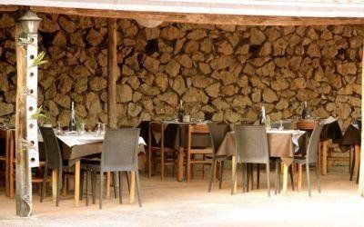 Zona pranzo rustica all'aperto con tavoli e sedie in legno e parete con legna da ardere accatastata.