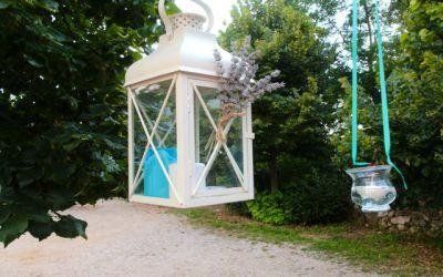 Una casetta per uccelli bianca a forma di lanterna, appesa tra gli alberi verdi lungo un sentiero del giardino.