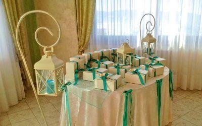 Tavolo dei regali con scatole decorate con nastri bianchi e verdi, lanterne e drappeggi di tessuto vicino a una finestra.