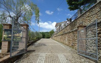 Un cancello di ferro aperto conduce a un vialetto in pietra in pendenza, accanto a un muro di pietra, sotto un cielo azzurro.