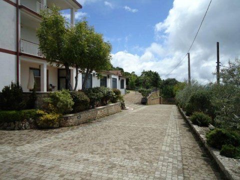 Viale d'accesso asfaltato accanto a edifici bianchi, arbusti e muri in pietra, sotto un cielo parzialmente nuvoloso.