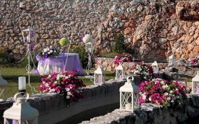 Tavolo nuziale all'aperto decorato con composizioni floreali viola e rosa e lanterne bianche accanto a un muro di pietra.