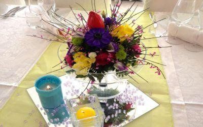 Candela blu e piccola composizione floreale su un tavolo con tovaglia gialla e dettagli rosa.
